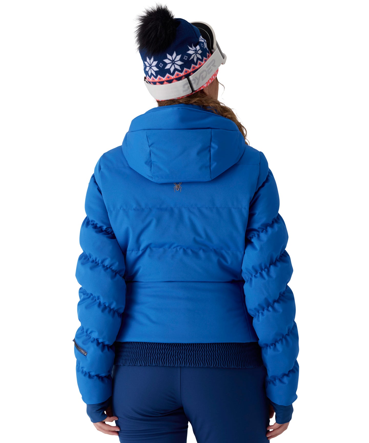 A woman viewed from behind wearing a bright blue puffer jacket, dark blue leggings, and a navy knit beanie with white snowflakes and a black pom-pom, against a light gray studio background.