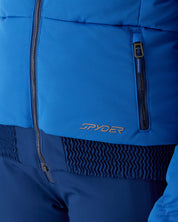 Close-up of a blue ski jacket showing the front zipper, a vertical zip pocket, and the Spyder logo embroidered in metallic gold on the lower front, paired with matching blue ski pants.