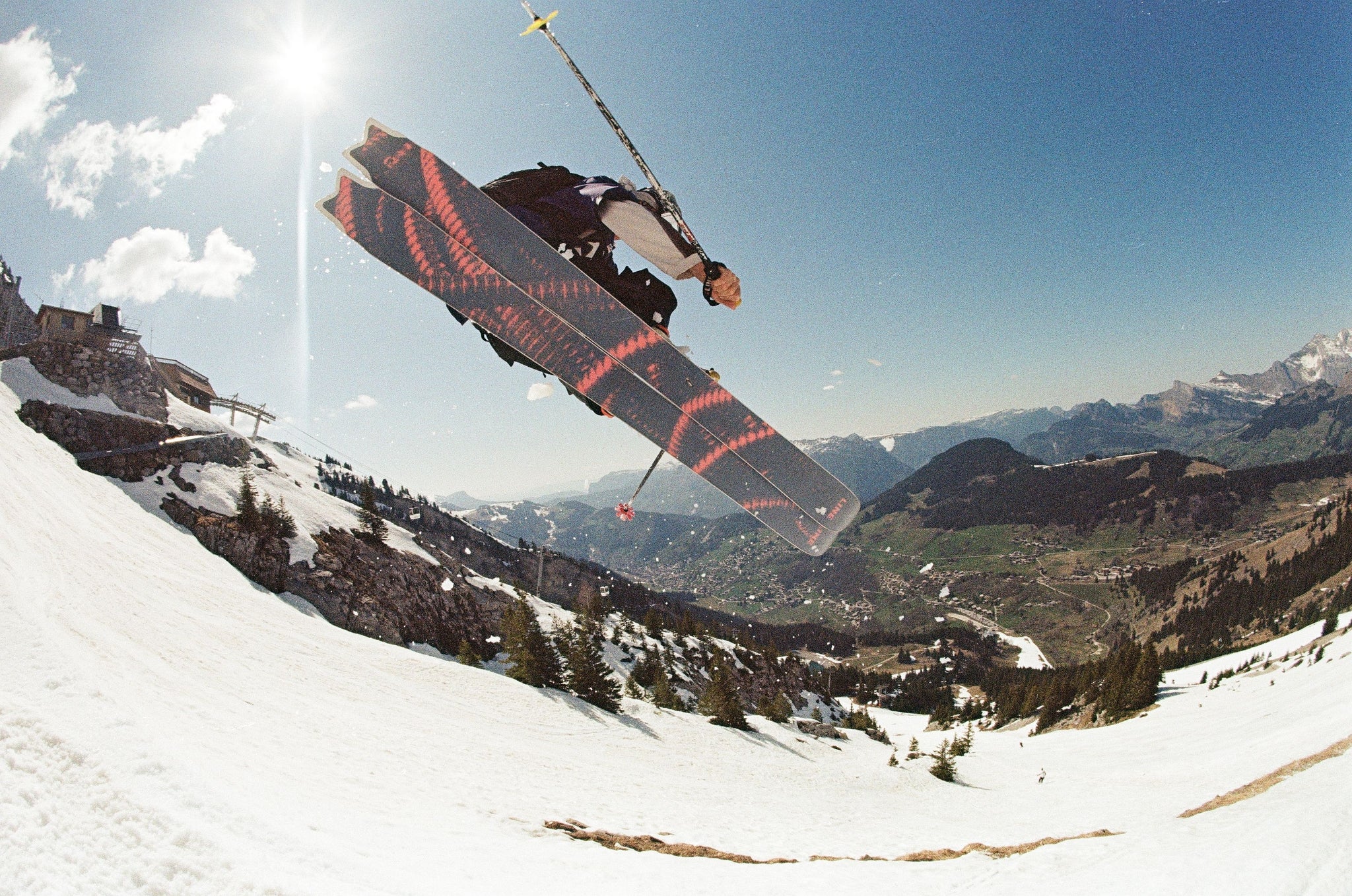 A skier mid-air performing a jump over a snowy mountain slope, with a valley and distant peaks under a bright blue sky with the sun shining.