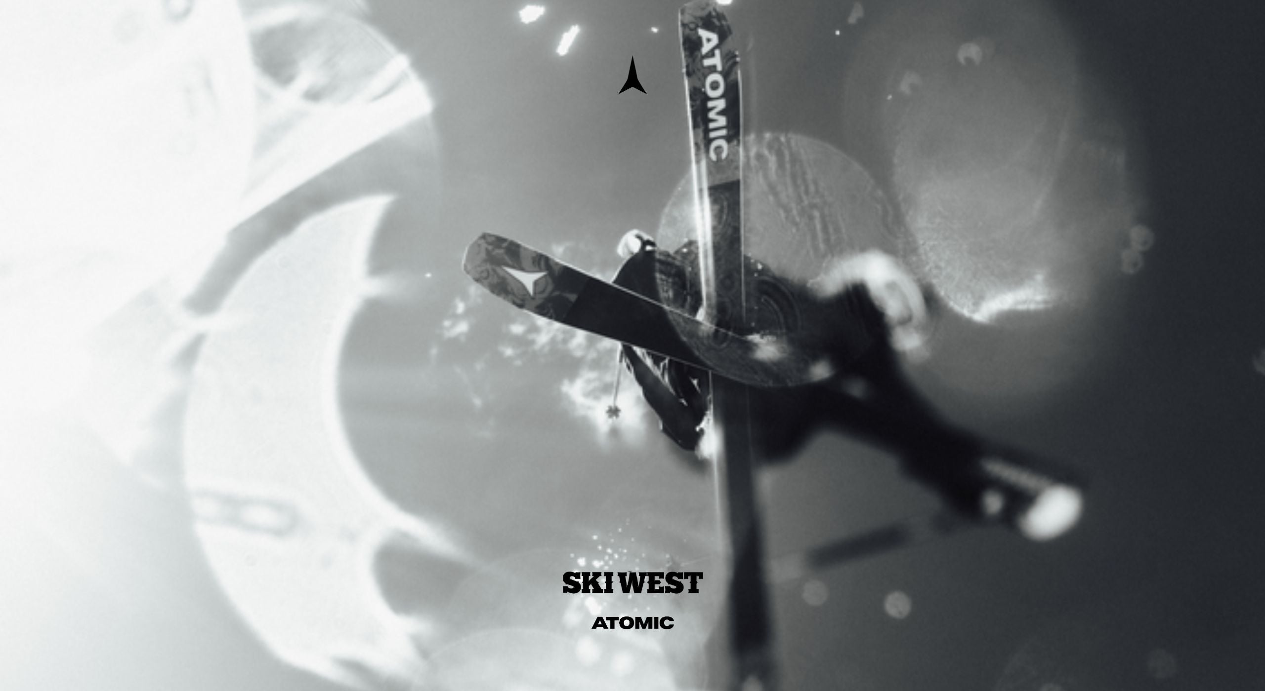 A monochrome promotional graphic featuring a skier in motion with a ski upright labeled 'ATOMIC' and 'SKI WEST ATOMIC' text near the bottom, set against a blurred abstract background.